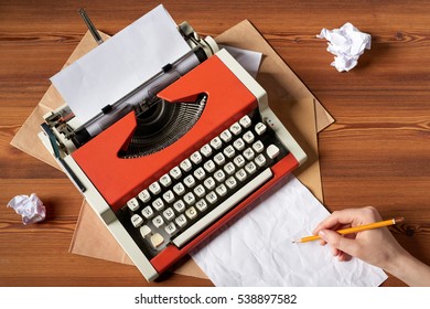 Top view of red vintage typewriter with white blank paper sheet with female hand holding pen, on wooden table