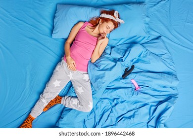 Top view of redhead European lying in comfortable bed wears casual pajama and headband looks at sex toys has pleased expression poses at cozy bedroom on bedclothes. People rest and intimacy concept