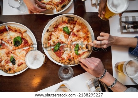 Top view restaurant table with Italian Margherita pizza and glasses of beer. Unrecognizable multiracial people's hands grabbing a sliced piece.