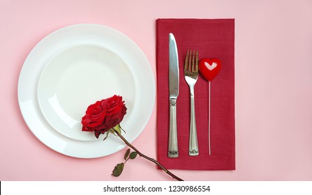 Top View Romantic Table Setting For Valentines Day Or Dinner Date Celebration Wedding. Table Setting With Red Rose Bud White Plates Fork And Knife On Red Tablecloth Pink Pastel Color Background. Space