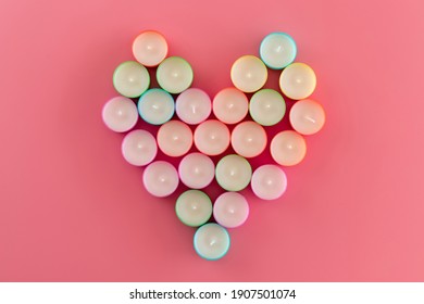 Top view of rows of unused multicolored wax tea light candles laid out in the shape of a heart  on pink background. Row of new water candles as background. A lot of candles pattern
