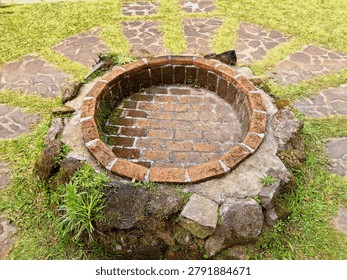 Top view of a rustic circular brick fire pit or decorative well in a green garden. Weathered stones, moss, and radiating flagstone pathways. Outdoor landscaping and backyard design concept.

