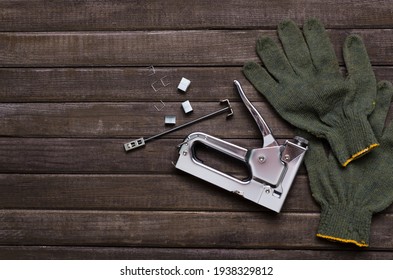 Top View Of Saple Gun, Staples And Green Protective Glove On The Brown Wooden Table.Empty Space