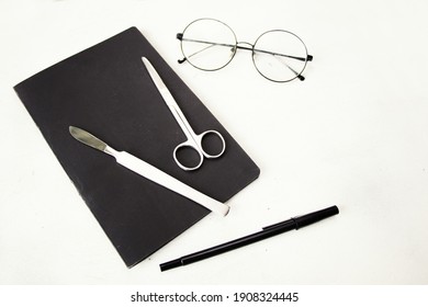 A Top View Of A Scalpel And Medical Scissors On A Notepad Next To Glasses And A Pen On A White Surface