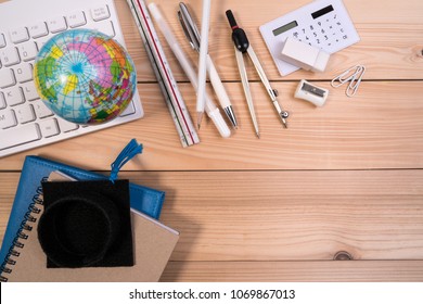 Top view of school accessories on wooden table desk with education supplies, blank note pad, calculator, pen, keyboard, earth globe model and graduation cap.
