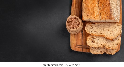 Top view of seeded bread loaf with sliced pieces and buckwheat on wooden board over black background with copy space