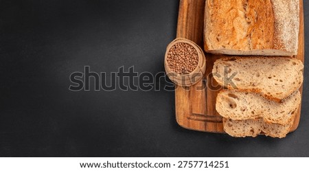 Top view of seeded bread loaf with sliced pieces and buckwheat on wooden board over black background with copy space