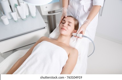Top View Of Serene Young Woman Getting Skin Rejuvenation Treatment. She Is Lying On Massage Table At Spa And Smiling. Cosmetologist Is Lifting Laser On Her Face