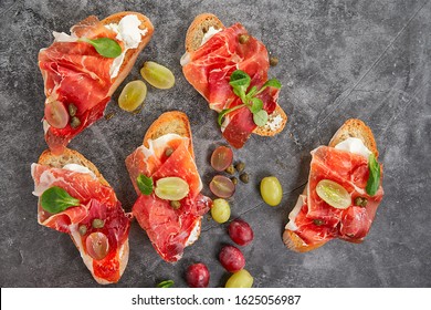 Top View Of Serrano Ham Tapas With White And Purple Grapes, Canons, Arugula. Capers And Honey On Black Background