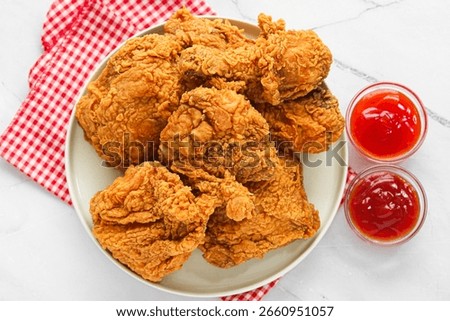 Top view of a serving of deep-fried chicken on a round white plate set on top of a red and white checkered cloth, paired with two small glass bowls of red dipping sauces on a marble surface.