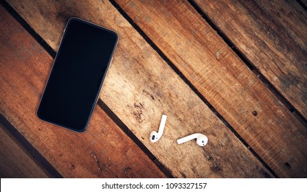 A top view of a set of wireless earbud headphones and mobile phone blank template lying on a rustic wooden table. 