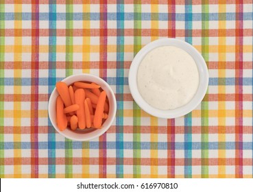 Top View Of Several Small Peeled Carrots In A Bowl With Ranch Dressing To The Side Atop A Colorful Cloth Patterned Place Mat.