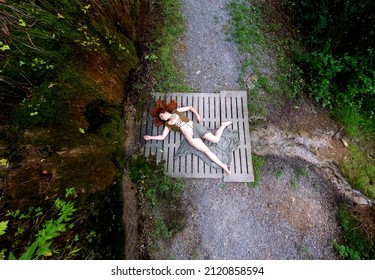 Top View Of Sexy, Young, Red-haired Woman Lies Dead On A Concrete Bridge Of A Forest Path On A Mossy Slope, After Accident Or Crime. What Happened ? Copy Space