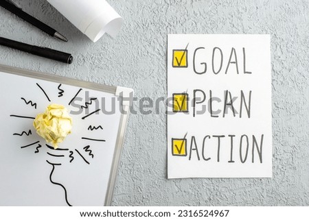 Top view of sheet with GOAL PLAN ACTION writing incompletely drawn thing and crushed wrapped paper on white desk on gray sand background