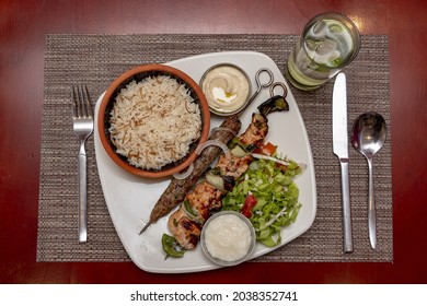 A Top View Of Shish Taouk, Kebab, Rice, And Cutlery On A White Plate On A Red Table