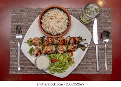 A Top View Of Shish Taouk, Rice, Sesame Paste, And Cutlery On A White Plate On A Red Table