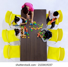 Top View Shot Of Asian Expertise Female Professional Lecturer Teacher Teach How To Play Colorful Brick Block Building Toy On Wood Table To Curious Eager Little Kinder Boy And Girls On Yellow Chairs.