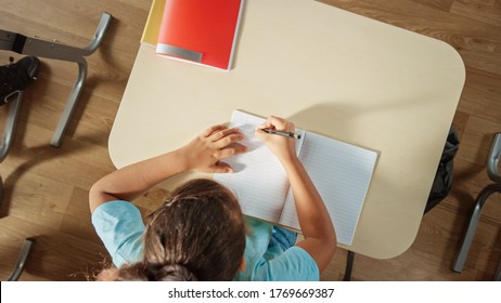 Top View Shot of Elementary School Classroom: Girl Sitting at the School Desk Working on Assignments in Exercise Notebooks. - Powered by Shutterstock