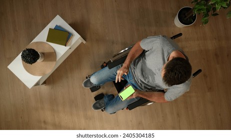 Top view shot a man with mobility impairment sitting on a wheelchair in a room, using a smartphone with an advertising area, workspace mock up on it. - Powered by Shutterstock