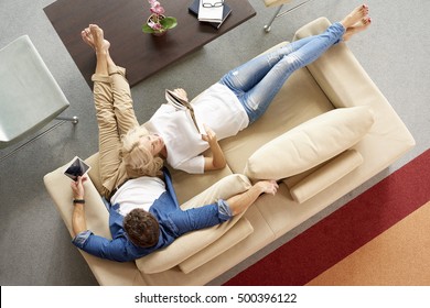 Top view shot of a middle aged couple relaxing together. Man with digital tablet sitting on couch while blonde women lying next to him and reading a book.