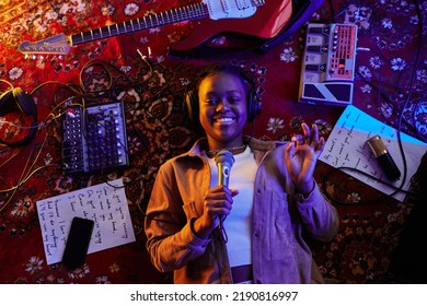 Top View Shot Of Smiling Black Woman Holding Microphone While Lying On Carpet In Purple Neon Light