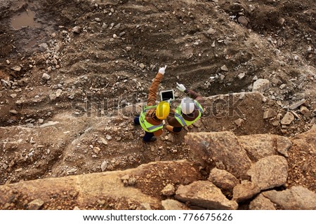 Top view shot of two industrial workers wearing reflective jackets standing on mining worksite outdoors using digital tablet, copy space