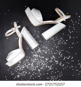 Top view of silver platform sandals with chunky block heels arranged diagonally on a black background scattered with white confetti dots. The shoes feature ankle straps and open toes.