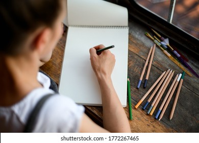 Top view sketchbook wooden table around which pencils lie   young woman draws in it 