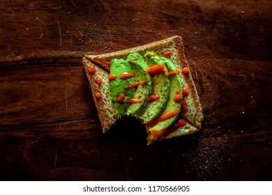 A Top View Of A Slice Of Bitten Toast Topped With Avocado And Sriracha Sauce