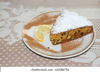 Top view of a slice of pumpkin walnut cake with powdered sugar on top, with a slice of lemon next to it as decoration, on a white plate.