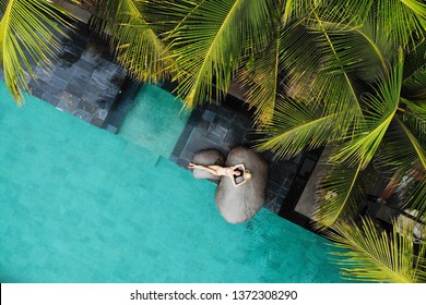Top View Of Slim Young Woman In Beige Bikini And Straw Hat Relaxing Near Luxury Swimming Pool And Palm Trees.Vacation Concept. Drone Photo