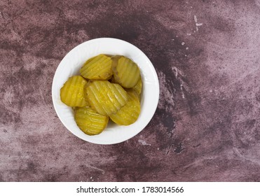 Top View Of A Small White Bowl Filled With Hamburger Dill Pickle Slices Offset On A Maroon Tabletop Illuminated With Natural Lighting.