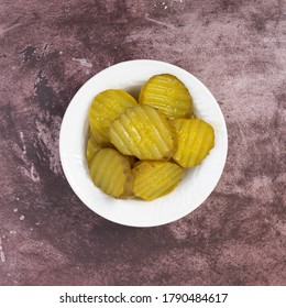 Top View Of A Small White Bowl Filled With Hamburger Dill Pickle Slices On A Maroon Tabletop Illuminated With Natural Lighting.