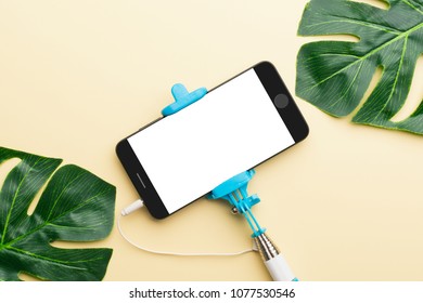 Top View Of Smartphone With White Empty Screen Connected With Selfie Stick And Composed With Green Monstera Leaves. 