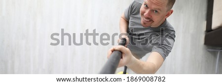 Top view of smiling middle-aged man cleaning apartment with grey vacuum cleaner. Cheerful person in grey shirt indoors. Renovation and construction site concept