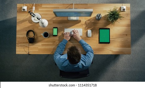 Top View Of A Software Engineer Working At His Desktop Computer Sitting In His Creative Office. Lying On The Table: Smartphone, Tablet Computer, Headphones.