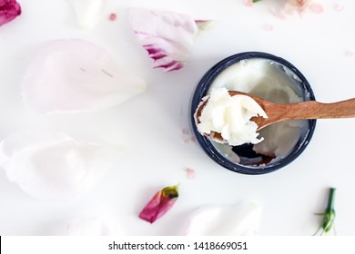 Top View Solid Shea Butter In A Black Jar With A Wooden Spoon On A White Background, Next To White And Pink Petals, Green Plant Twigs. Copy Space.