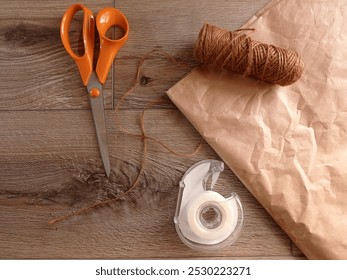 Top view of some packaging material and tools (scissors, adhesive tape, jute string and recycled paper) on a wooden table.