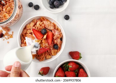 top view of someone's pouring fresh milk into a bowl of strawberry corn flakes cereal and blueberries for simple and fast breakfast on the table  - Powered by Shutterstock