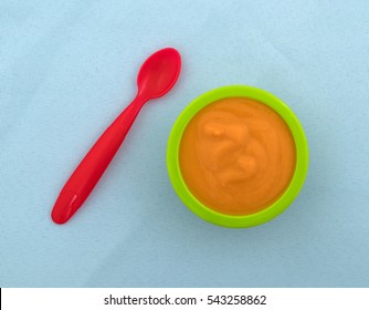 Top View Of A Spoon With A Serving Of Banana Carrot And Mango Baby Food In A Green Bowl Atop A Wrinkled Blue Tablecloth.