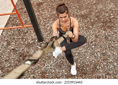 Top view of sportswoman doing sit ups for ass muscles with suspension straps on sports ground. Concentrated adult caucasian woman with tattoos wearing sportswear. Modern healthy and sports lifestyle