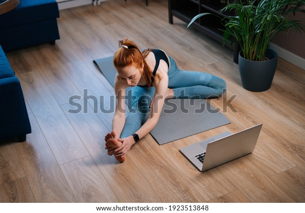 Top view of sporty redhead young woman\
working out, doing stretching exercise on yoga mat while watching\
fitness video online on laptop. Concept of sports training\
red-haired lady during\
quarantine.