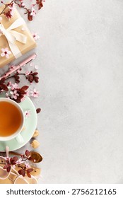 Top view spring composition with tea cup on mint saucer, cherry blossoms, wrapped gift, pen, glasses, and golden spoon on a light gray background. Minimal lifestyle flat lay with wide copy space.