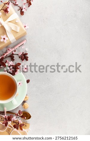 Top view spring composition with tea cup on mint saucer, cherry blossoms, wrapped gift, pen, glasses, and golden spoon on a light gray background. Minimal lifestyle flat lay with wide copy space.