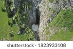 Top view of steep mountain cliffs and an ice cave. An avalanche descended into the gorge, and a snow tunnel (ice grotto) was formed. Mountainous terrain, spruce trees and green grass grow.