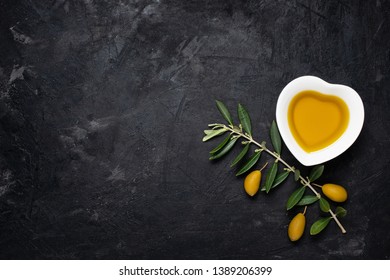Top view of still life .In the ceramic bowl, in the shape of a heart, extra virgin olive oil, on the rough black background a twig with green olives. Free space for text