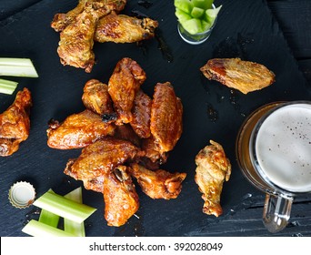 Top View Of Stone With Crispy Delicious Buffalo Chicken Wings With Hot Spicy Sauce. Black Wood Background With Celery Sticks And Beer On It