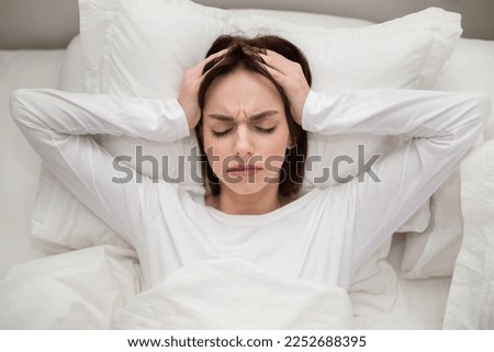 Top view of stressed sad irritated young brunette woman wearing white pajamas lying in bed at home, touching head with both hands, feeling tired after sleepless night. Insomnia, sleep disorder conept