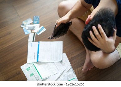 Top view of stressed young Asian man holding empty wallet and thinking about  finding money to pay credit card debt and all bills. He is holding head by another hand. Financial problem concept.