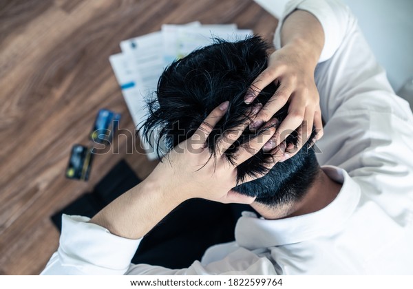 Top view of stressed young Asian man sitting and\
hugging knees, trying to find money to pay credit card debt and all\
loan bills. Financial problem from coronavirus or covid19 outbreak\
crisis concept.